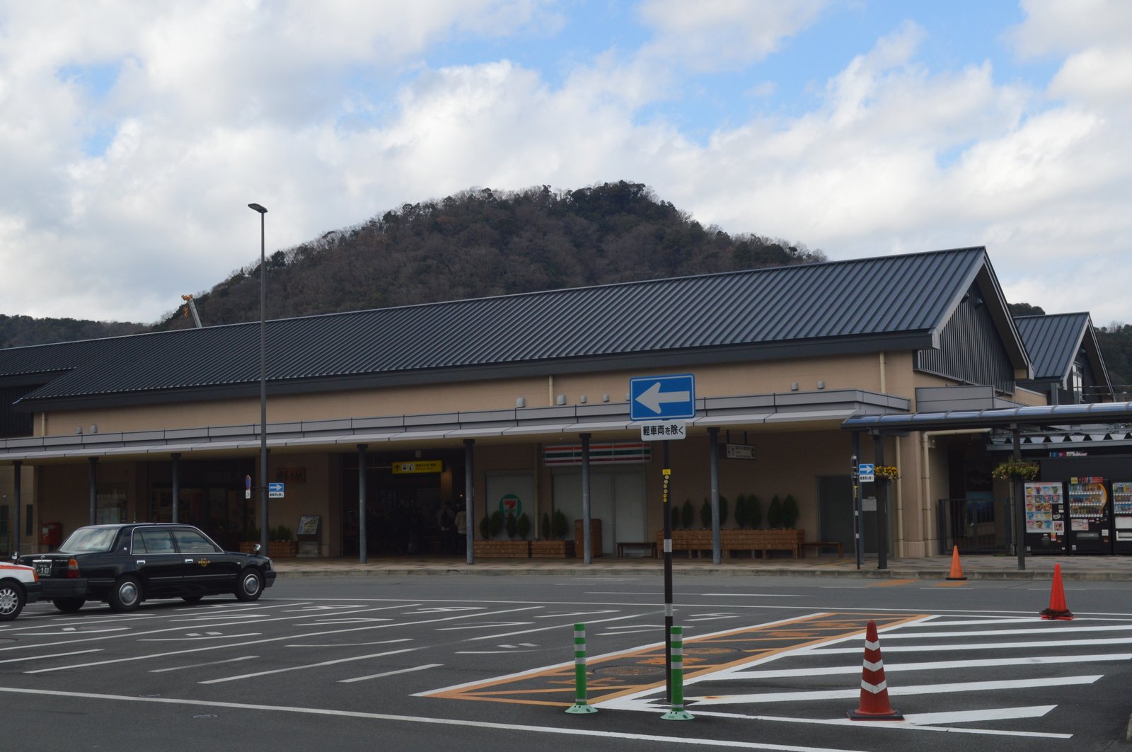 黒い瓦屋根の修善寺駅の駅舎。背後に木々の茂る山が見え、手前のロータリーにタクシーが停まっている