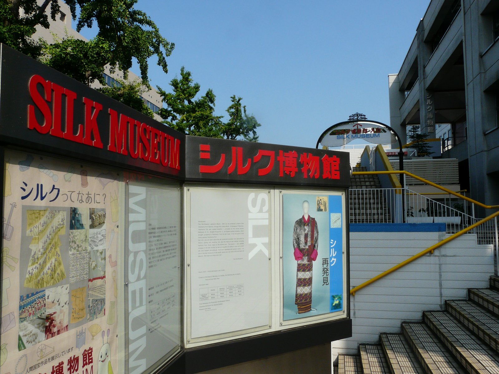 赤い「SILK MUSEUM」「シルク博物館」の文字が掲げられた入口の看板。下の掲示板には「シルクってなあに？」の解説と絹の衣装の写真があり、右手に階段が見える
