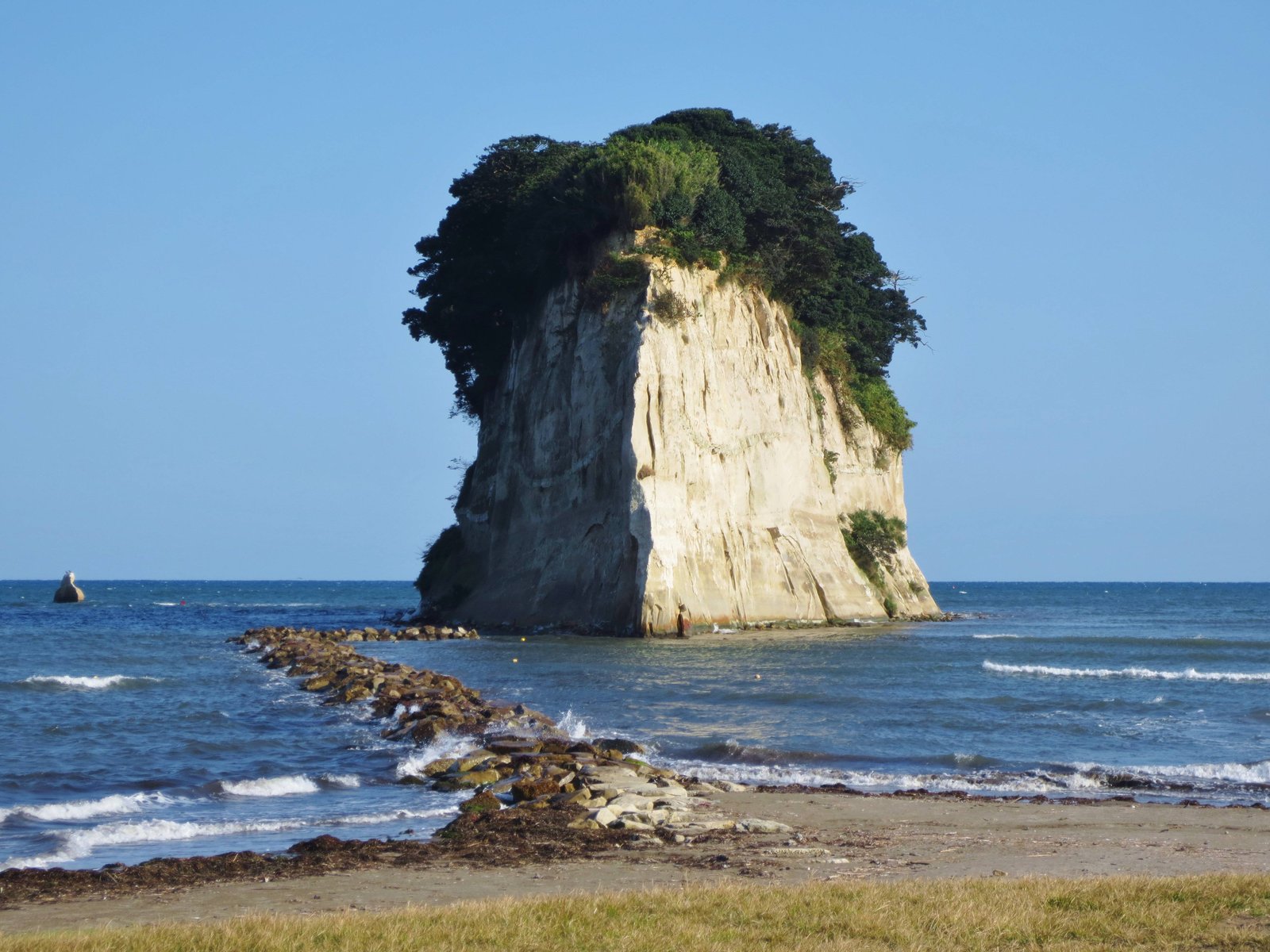 海岸から沖へ延びる石の並びの先に立つ見附島。白い岩の上を木々が覆っている