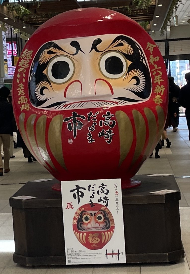高崎駅の構内に置かれた大きな赤いだるま。胴に「高崎だるま市」と書かれ、台には市のポスターが貼られている