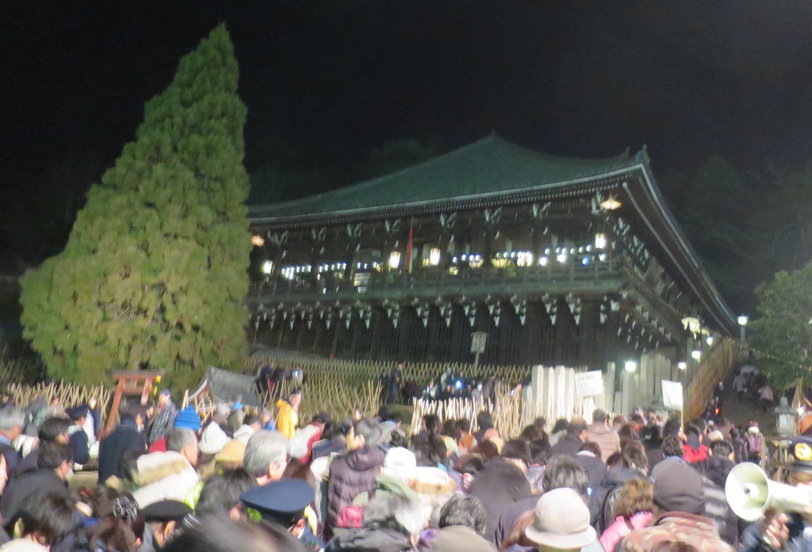 夜の二月堂。舞台の明かりがともり、下の広場に大勢の人が座って集まっている。手前に竹の柵