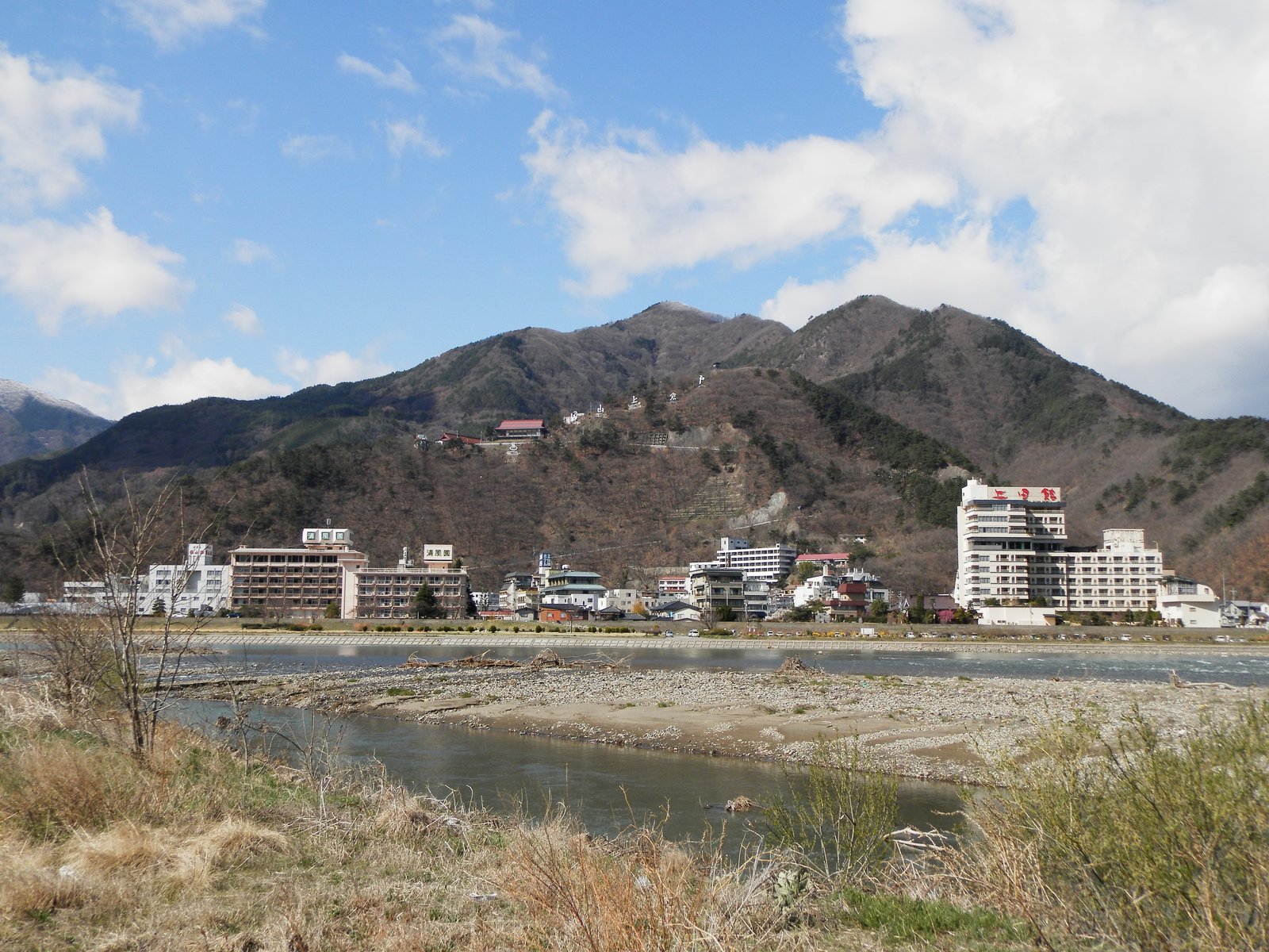千曲川の河原の向こうに、戸倉上山田温泉の旅館の建物が横に並び、背後に冬枯れの山が連なって、手前には川の流れと枯れ草が見える