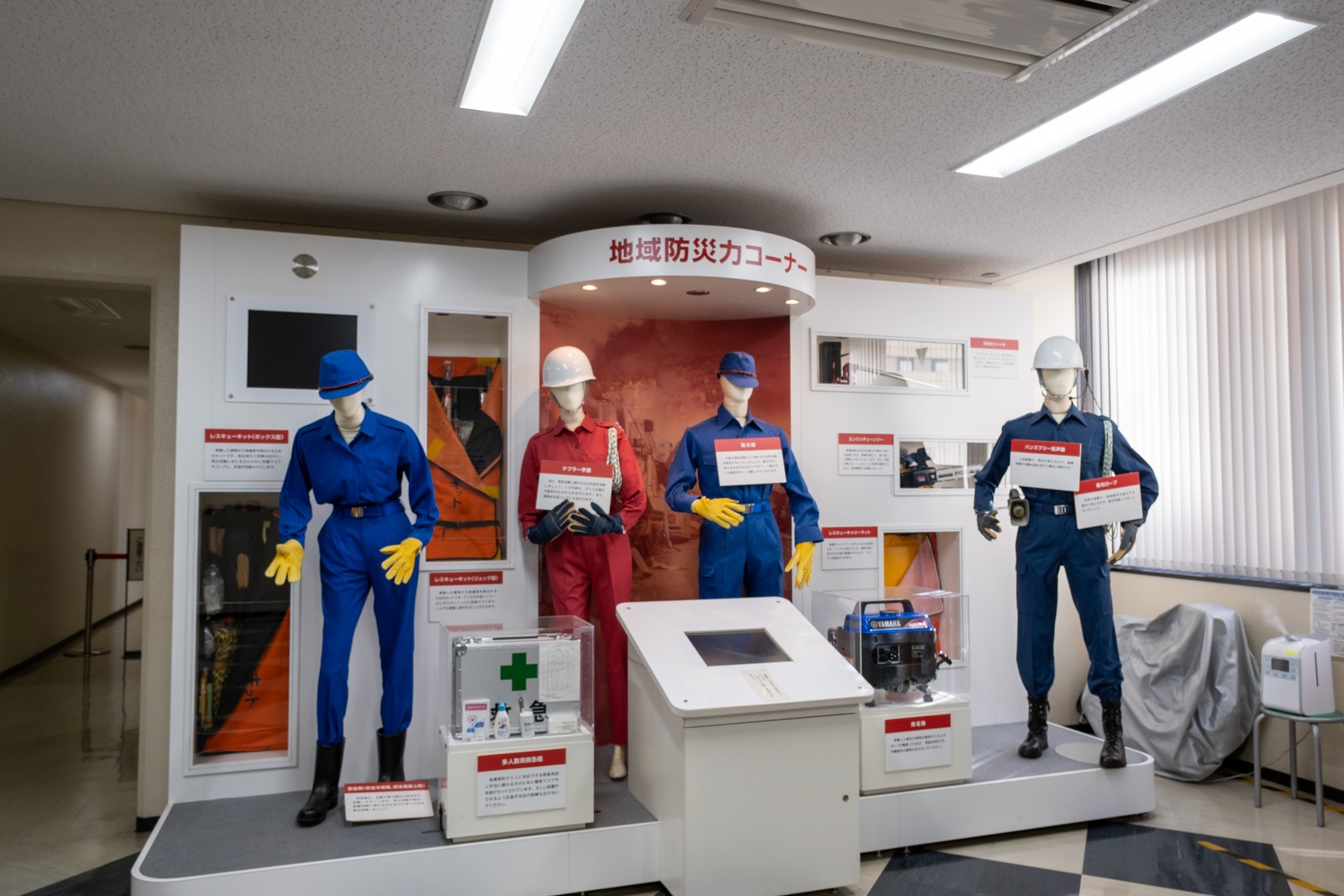 池袋防災館「地域防災力コーナー」の展示。消防団や防災の装備を着けたマネキン4体と、レスキューキット・多人数用救急箱・発電機などの資機材が並ぶ