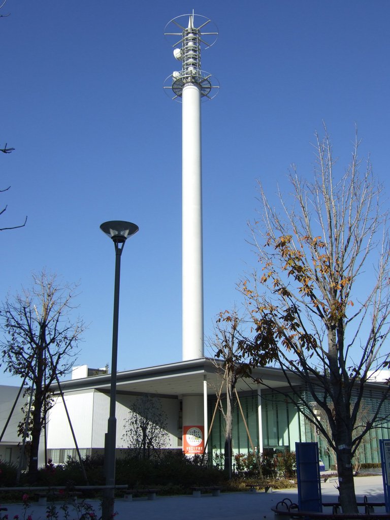 青空の下、白く細長い塔がそびえるそなエリア東京の建物。低い建物の前に街灯と葉の残る木が並ぶ