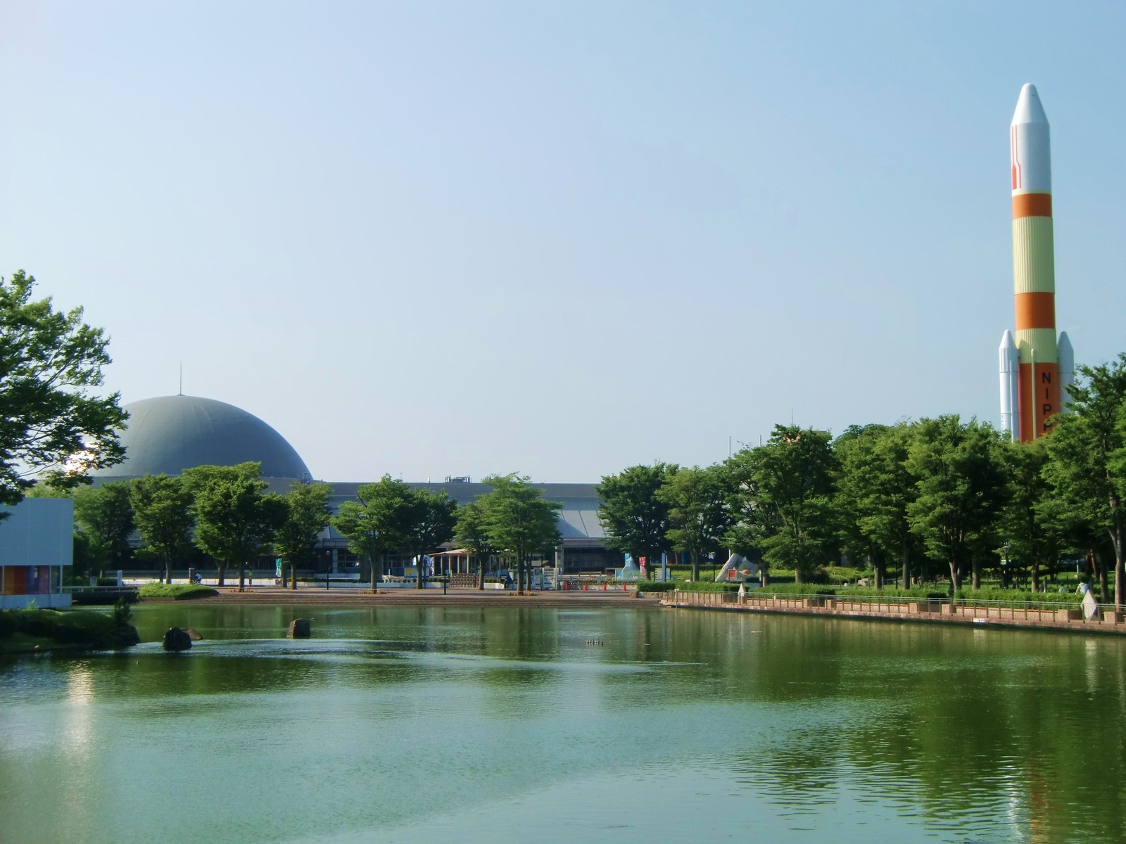 池の向こうに立つつくばエキスポセンター。灰色のドームを載せた低い建物が木立の奥に見え、右手に白とオレンジのH-IIロケットの模型がそびえる