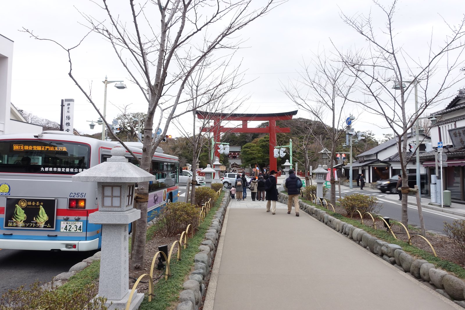 石垣で一段高くした参道の段葛。両側に石灯籠と葉を落とした桜の木が並び、正面に朱色の大きな鳥居、その奥に八幡宮の社殿が見える。左の車道に路線バス