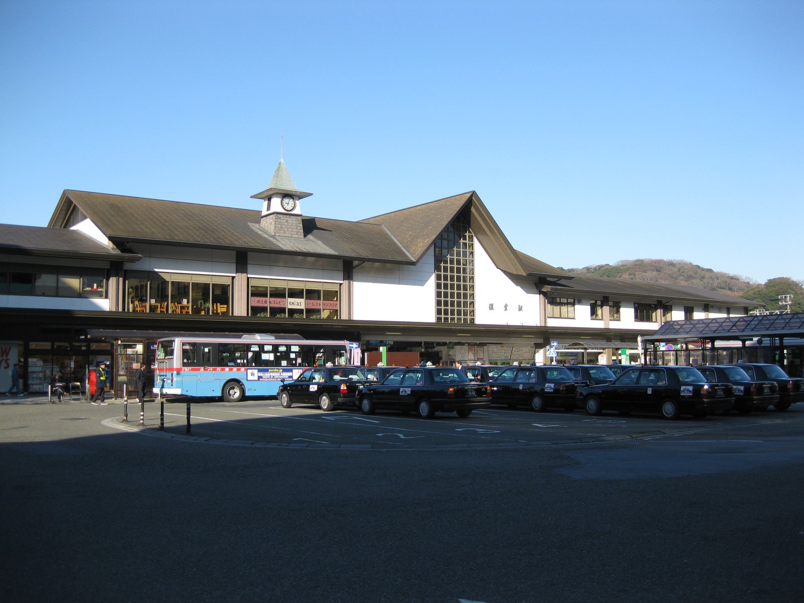 時計塔のある三角屋根のJR鎌倉駅の駅舎。駅前の広場に黒いタクシーが並び、水色の路線バスが停まっている