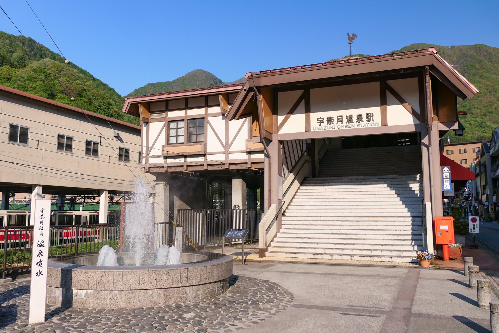 山を背にした宇奈月温泉駅の駅舎。切妻屋根の入り口に「宇奈月温泉駅」の看板があり、手前の石組みの噴水が水を上げている