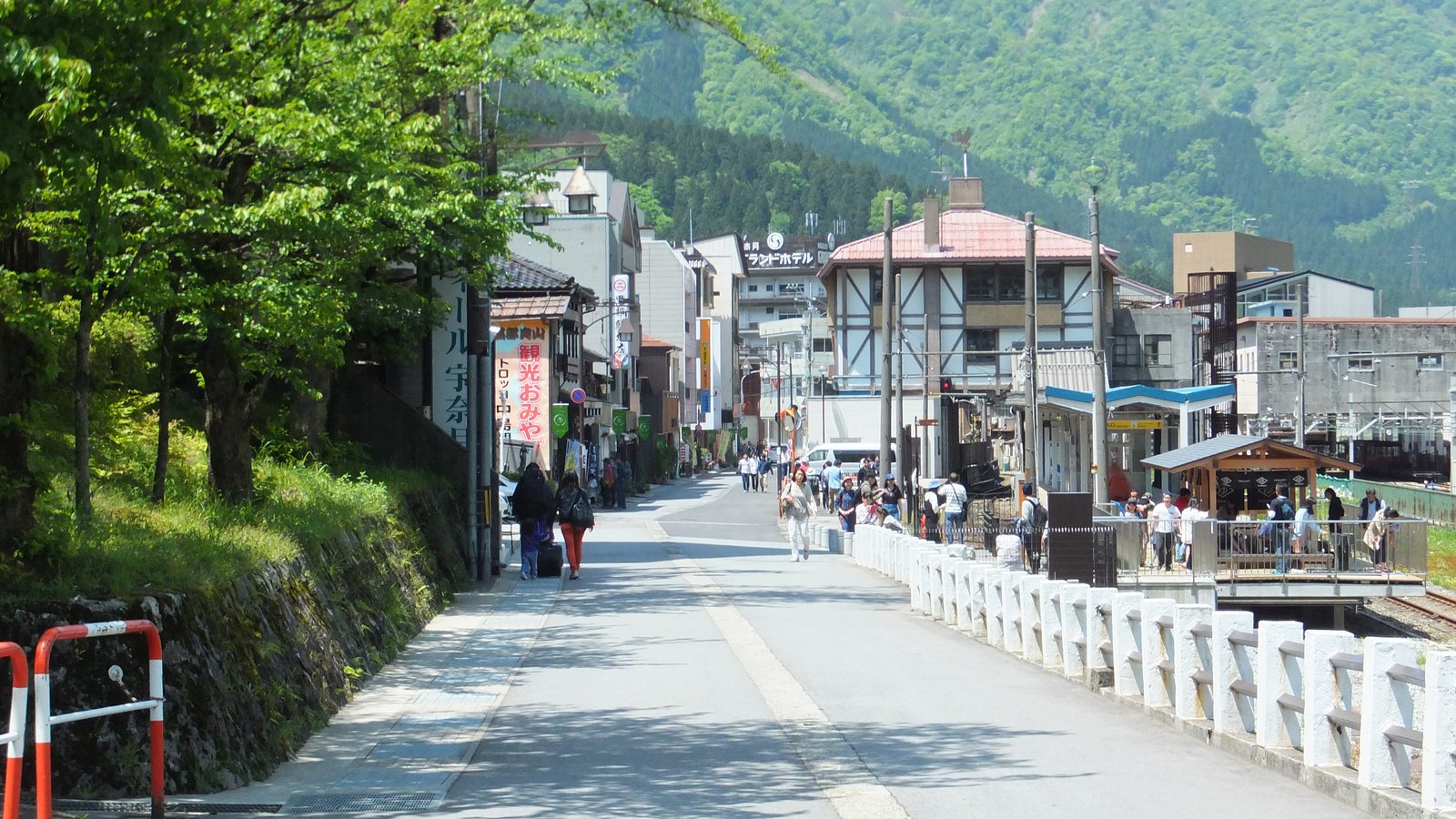 新緑の山を背にした宇奈月温泉の通り。左に木立と土産物店ののぼり、奥にホテルや旅館の建物が並び、右手の白い柵の向こうに線路と駅の屋根が見える