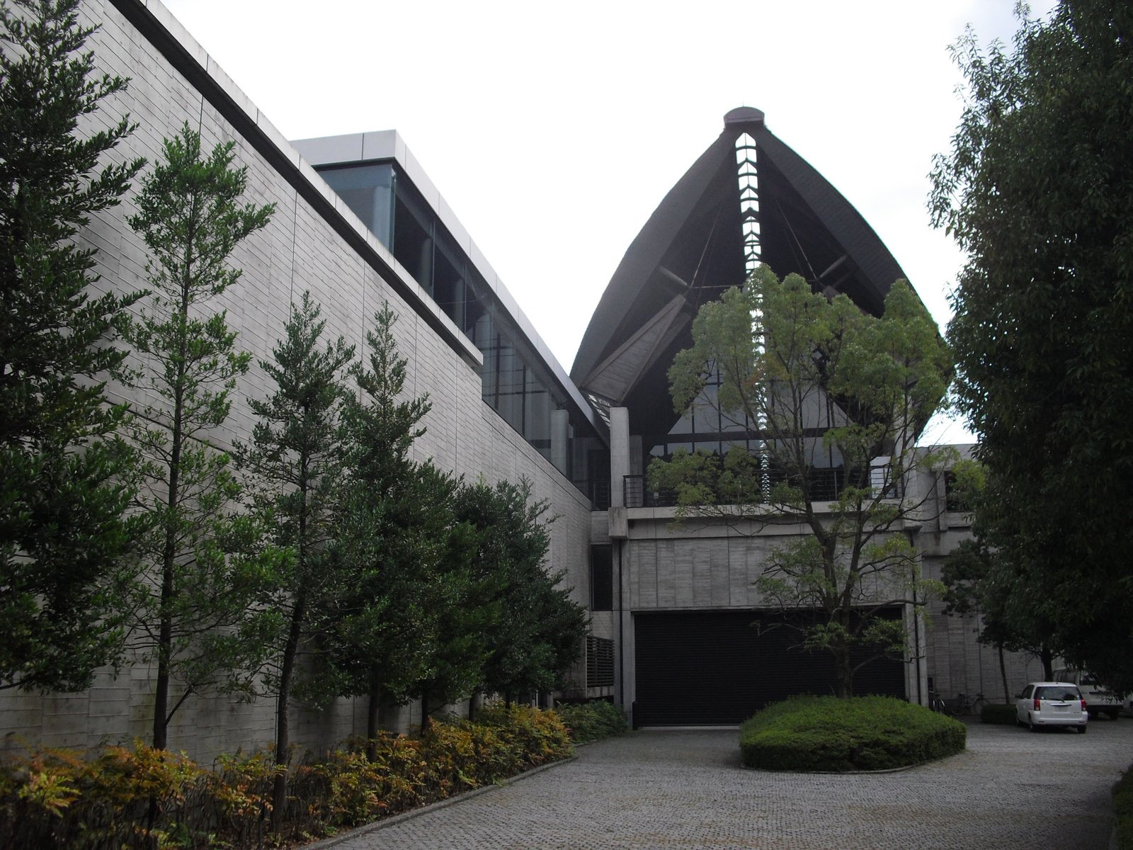 愛媛県歴史文化博物館の外観。打ち放しコンクリートの長い壁の奥に、ガラスを組んだ三角形の大きな屋根の建物が立ち、前庭に丸く刈った植え込みと木がある