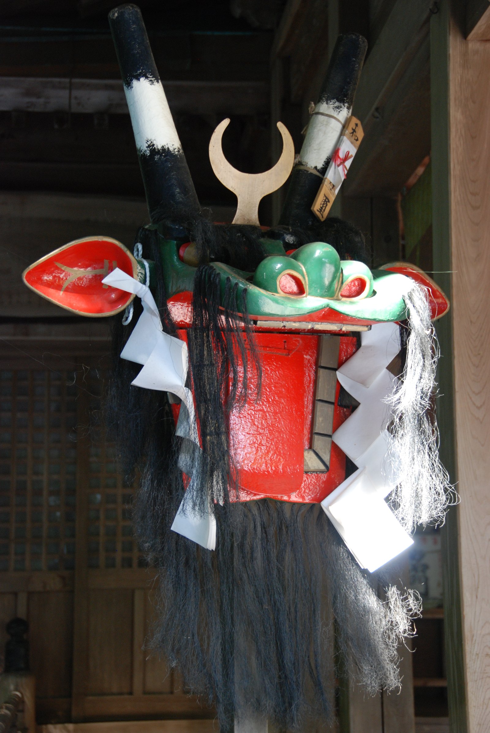 和霊神社に置かれた牛鬼の頭。赤く塗った顔に緑色の鼻、白い紙の歯と黒い毛が付き、上に二股に分かれた金色の飾りと赤い宝珠、後ろに黒と白の縞の首の棒が伸びる