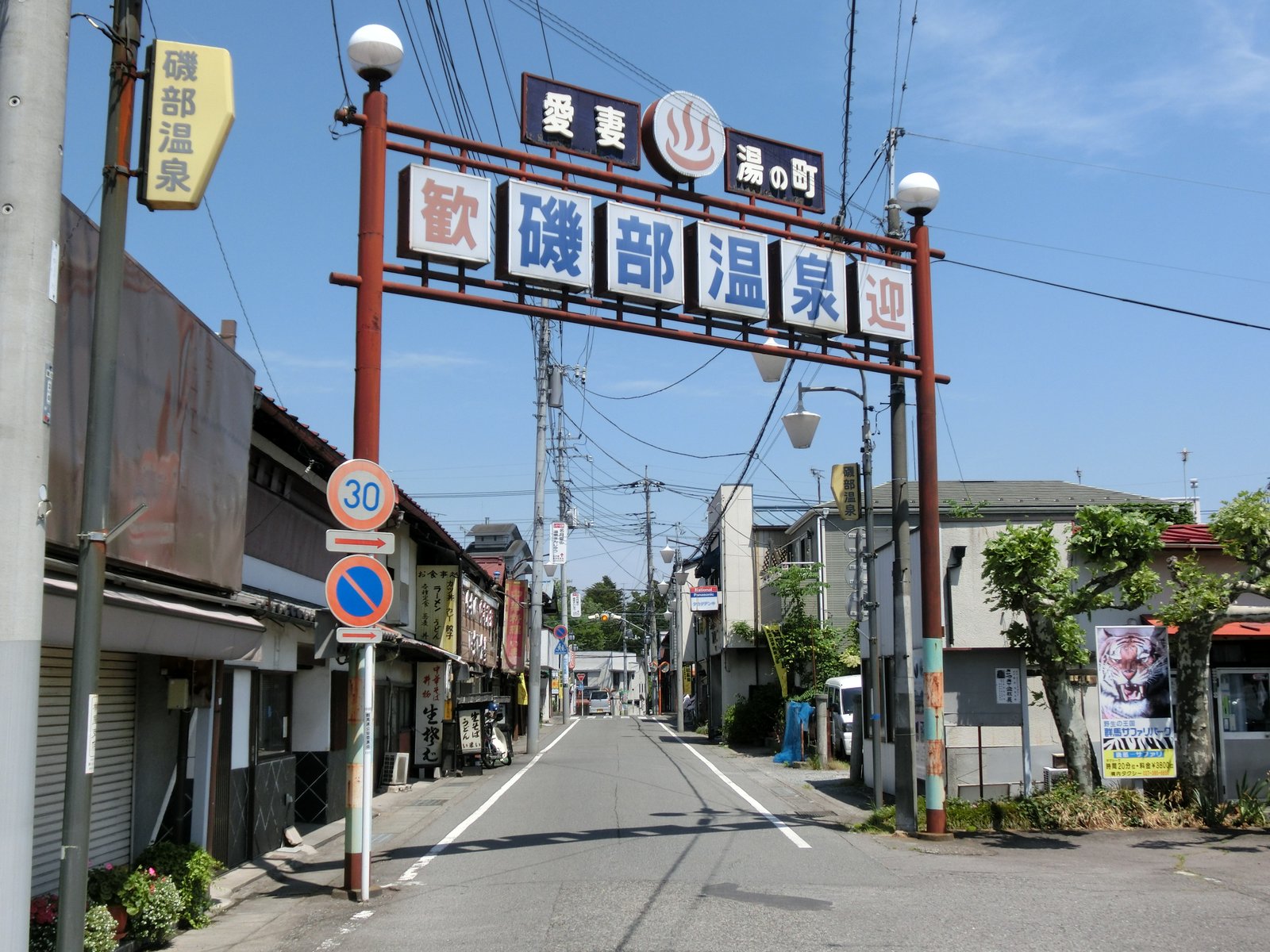 「愛妻 湯の町」「歓迎 磯部温泉」と書かれた赤い柱の看板アーチが立つ通り。両側に店や家が並び、まっすぐな道が奥へ続く