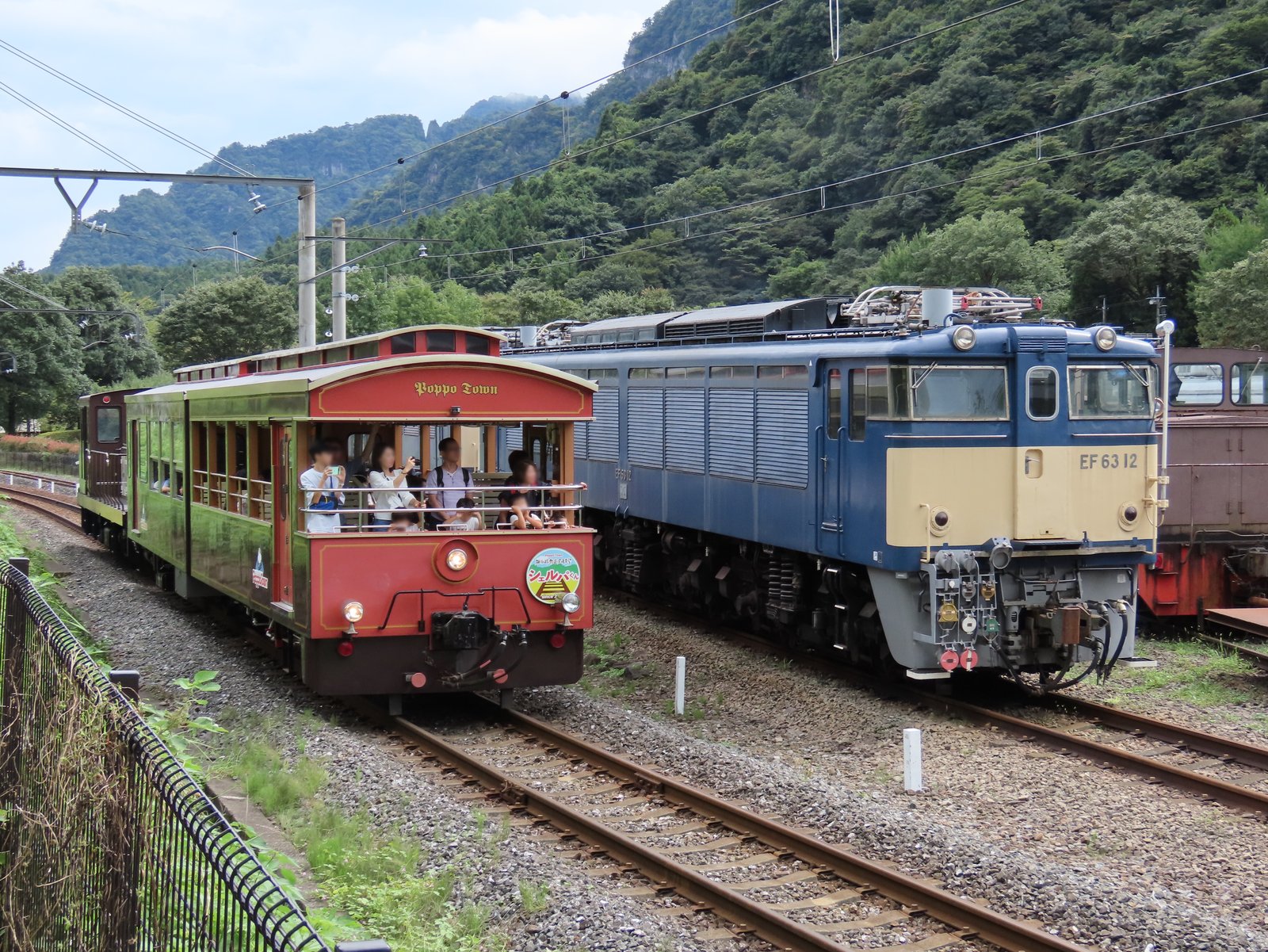 碓氷峠鉄道文化むらで、乗客を乗せた赤い屋根のオープン型トロッコ列車「シェルパくん」が、青とクリーム色の電気機関車EF63形12号機の横を走る。背後は緑に覆われた山