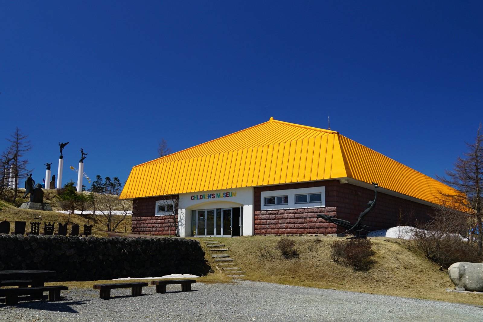 黄色い大きな屋根と赤茶色の石張りの壁を持つこども美術館。入口の上に「CHILDREN'S MUSEUM」の文字があり、手前に砂利の広場と木のベンチがある