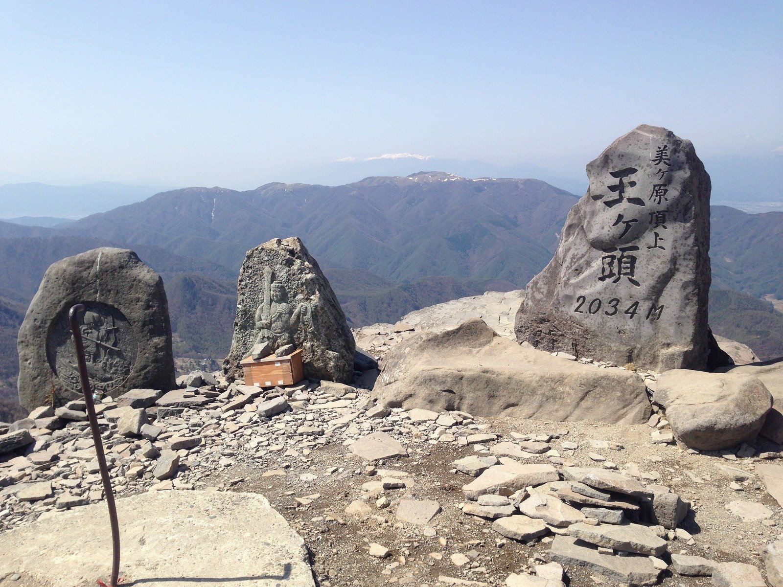 岩だらけの山頂に立つ、「美ヶ原頂上 王ヶ頭 2034M」と刻まれた石碑。左に石仏を彫った石が並び、奥に山並みが広がる