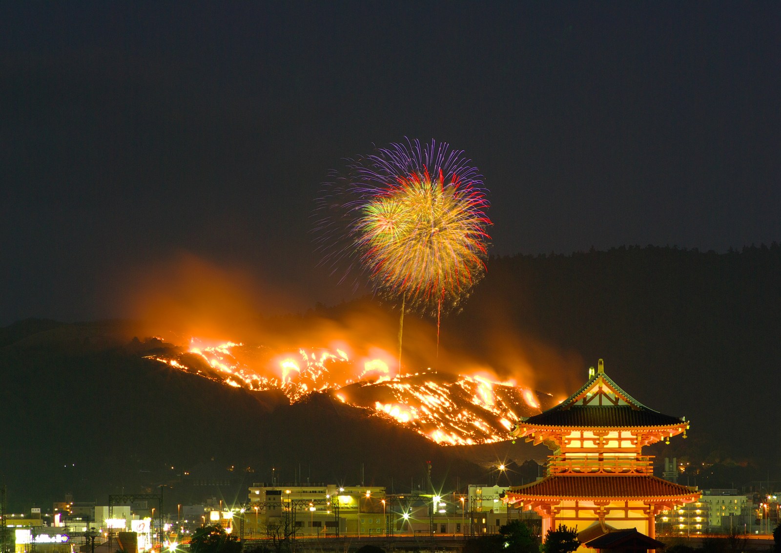 夜の若草山焼き。山肌が帯のように燃え、その上に花火が開いている。手前に朱塗りの二重の門が照らされて立つ