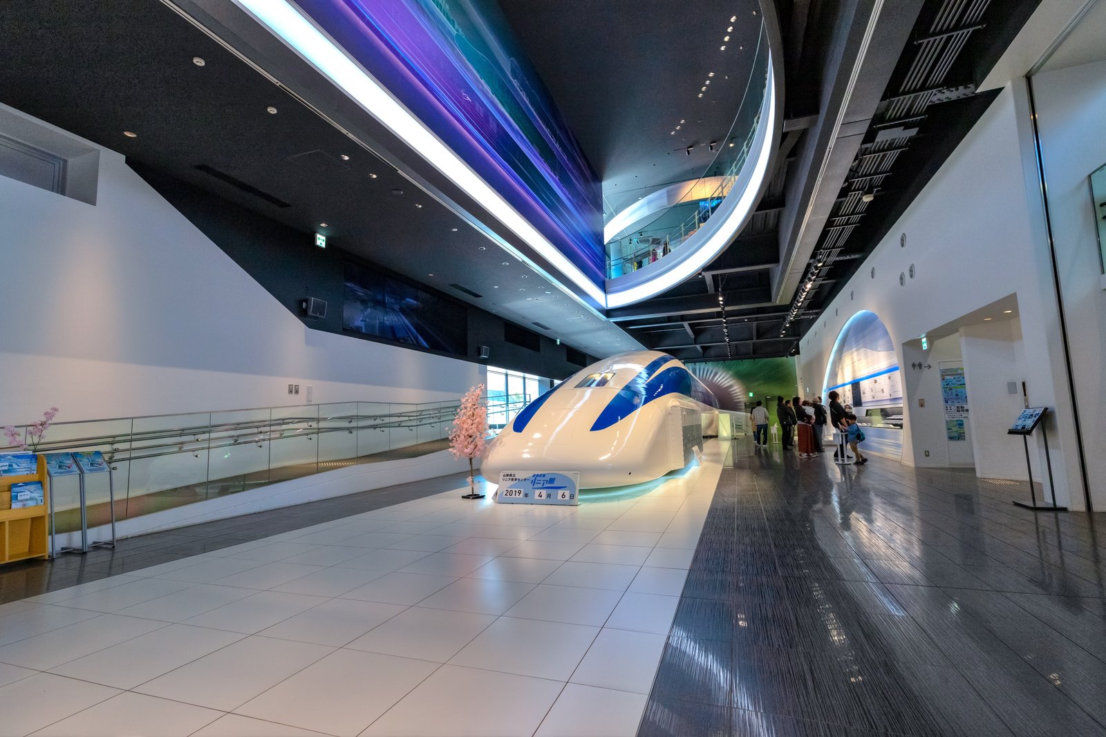 山梨県立リニア見学センターの館内に展示された超電導リニアの先頭車両。天井の曲面照明の下で、来館者が展示を見ている