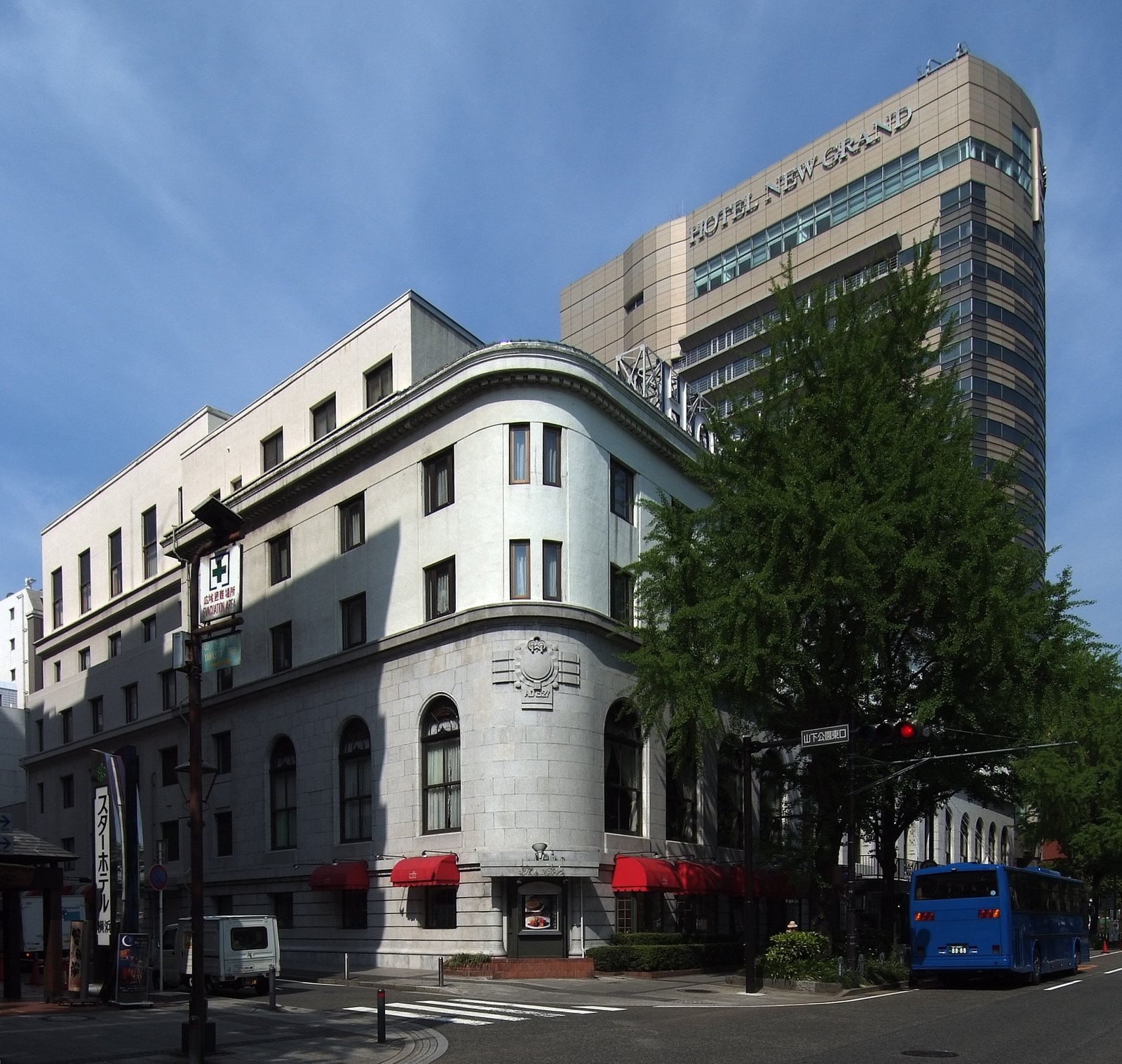 ホテルニューグランド本館。角の丸い白い石造りの建物で、1 階の窓に赤いひさしが付き、奥に HOTEL NEW GRAND の文字を掲げた高い建物が立つ