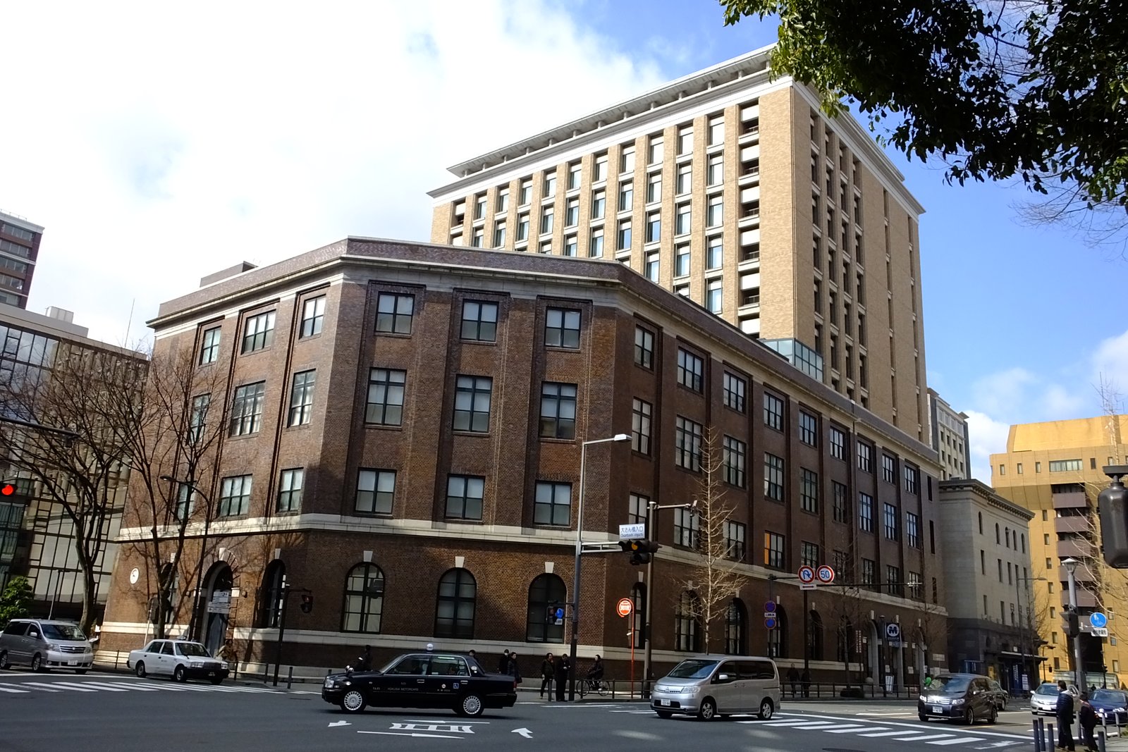 日本大通りの交差点から見た、濃い茶色のタイルを張った4階建ての旧横浜市外電話局。1階にアーチ形の窓が並び、奥に高層ビルが建つ