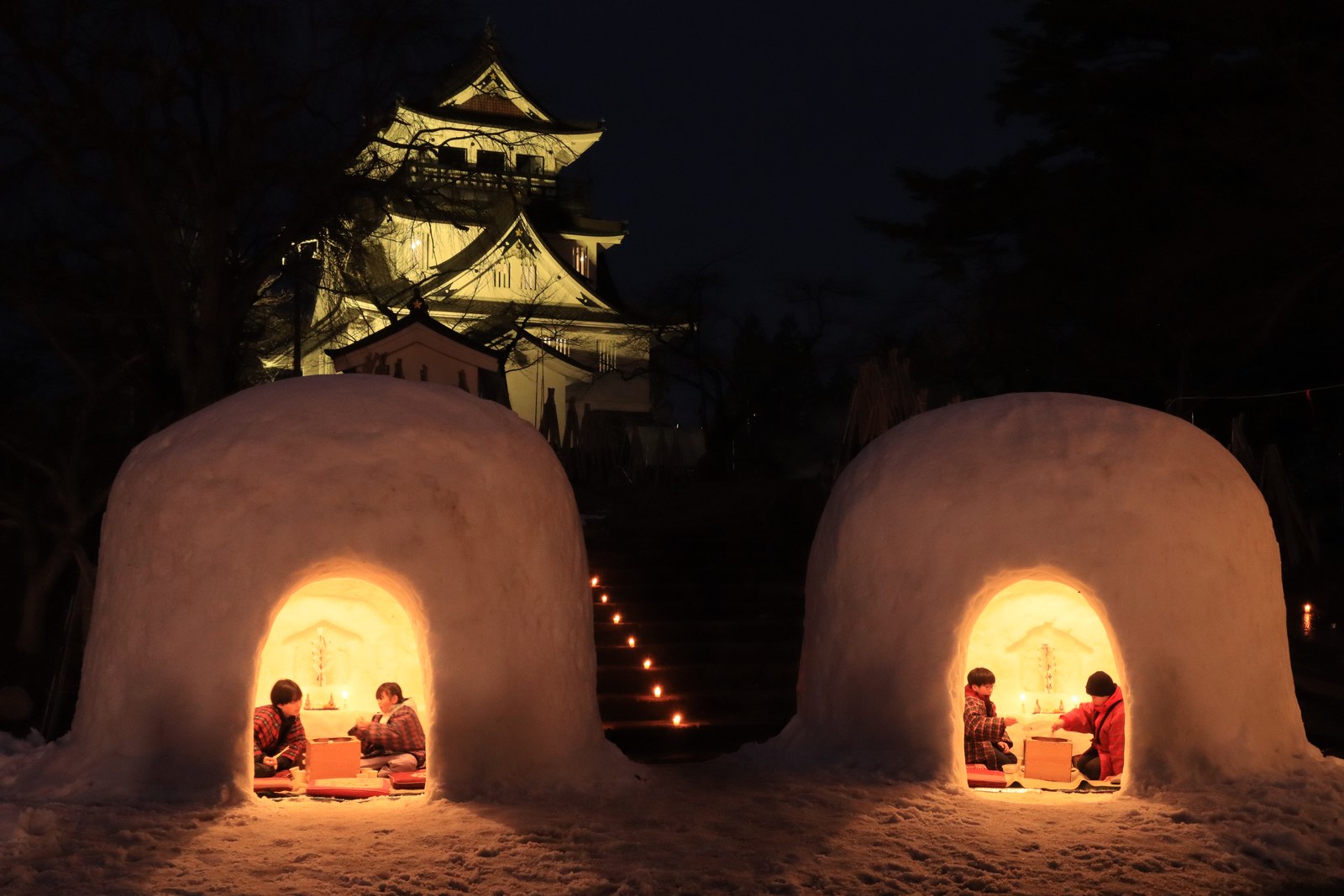 夕暮れの横手公園。灯りのともったかまくらが 2 基並び、奥の高台に横手城の天守がライトアップされている