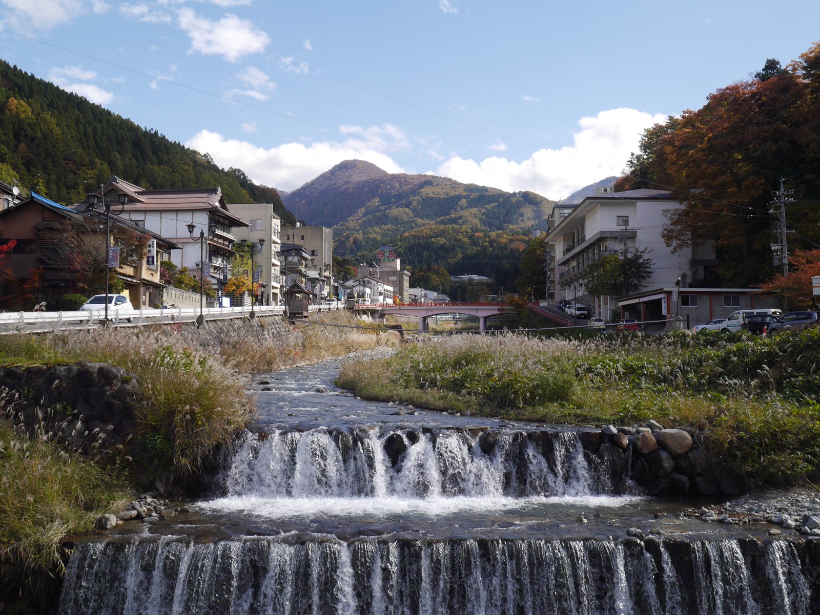 小さな堰を水が流れ落ちる川の両岸に、旅館が建ち並ぶ渋温泉。奥に赤い橋と山があり、手前の土手にススキが生えている。