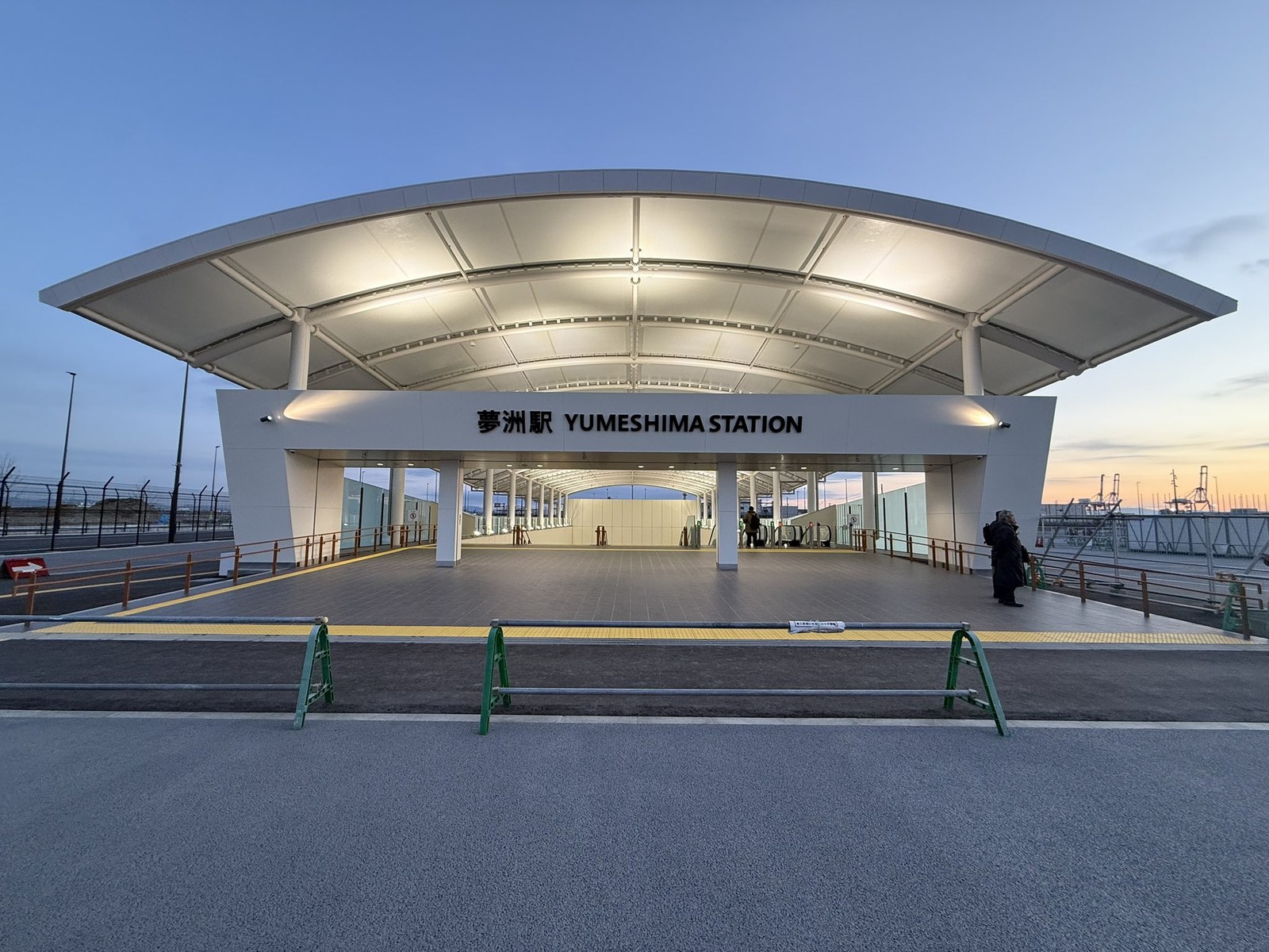 夕暮れの夢洲駅の出入口。白い大屋根の下に「夢洲駅 YUMESHIMA STATION」の表示があり、奥にコンテナ埠頭のクレーンが見える