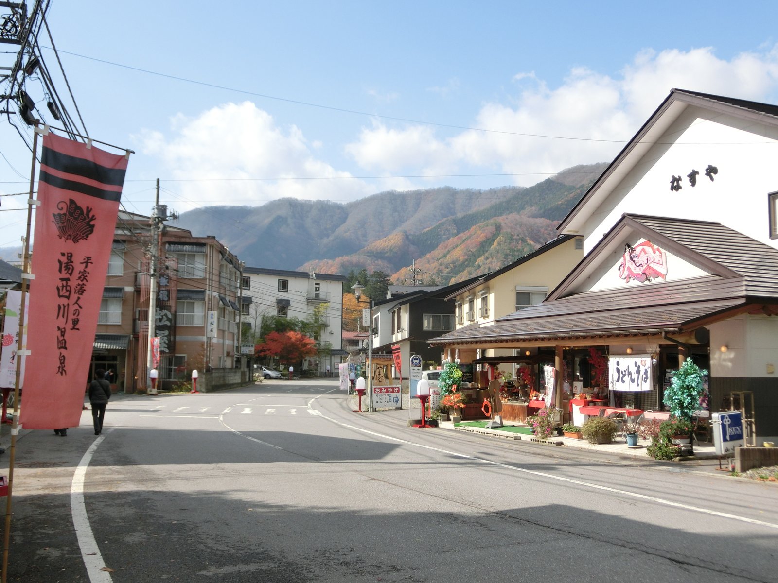 「平家落人の里 湯西川温泉」と書かれた赤いのぼりが立つ通り。右に土産物店とうどん・そばの店、左に宿の建物、奥に紅葉した山が見える