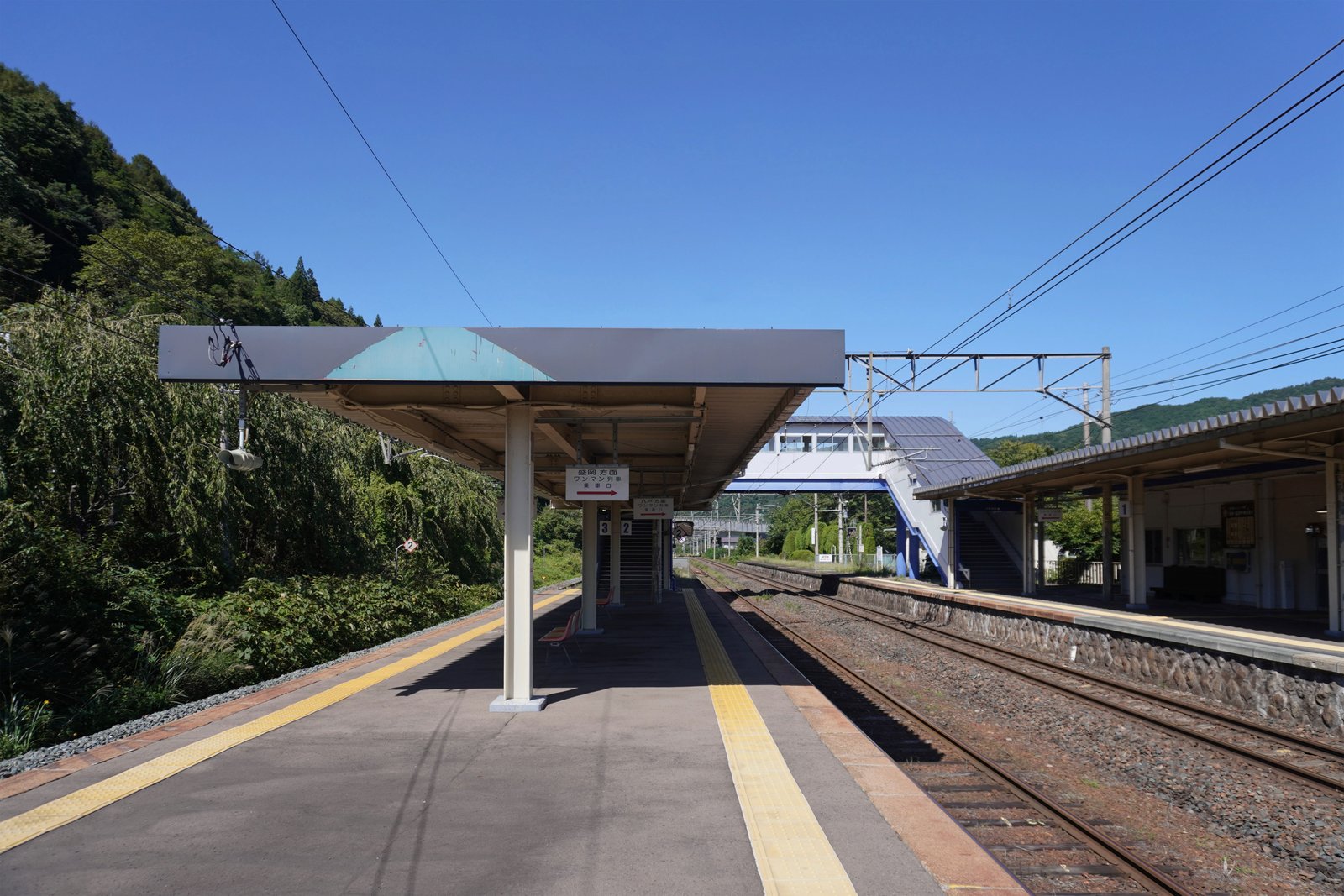 金田一温泉駅のホーム。屋根の下に「盛岡方面 ワンマン列車乗車口」の案内板が下がり、線路の向こうに青い跨線橋と駅舎、背後に緑の山が見える