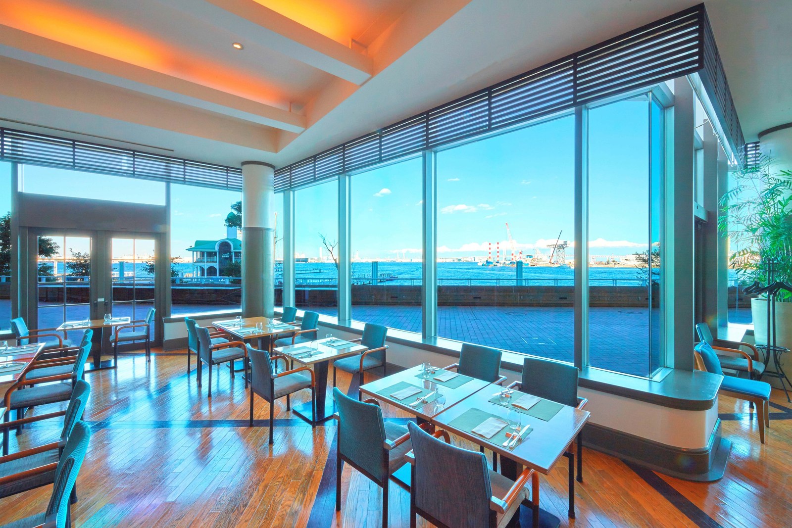 昼の1階レストランの店内。大きな窓の向こうにテラスと海、緑の屋根の建物が見え、テーブルと椅子が並ぶ