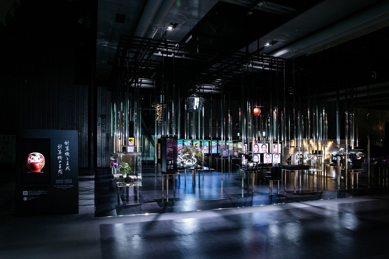 薄暗い展示室の中央に、細い金属の柱を何本も立てた展示。柱の間に蝶や草花のような形が浮かび、奥の壁に映像が映っている