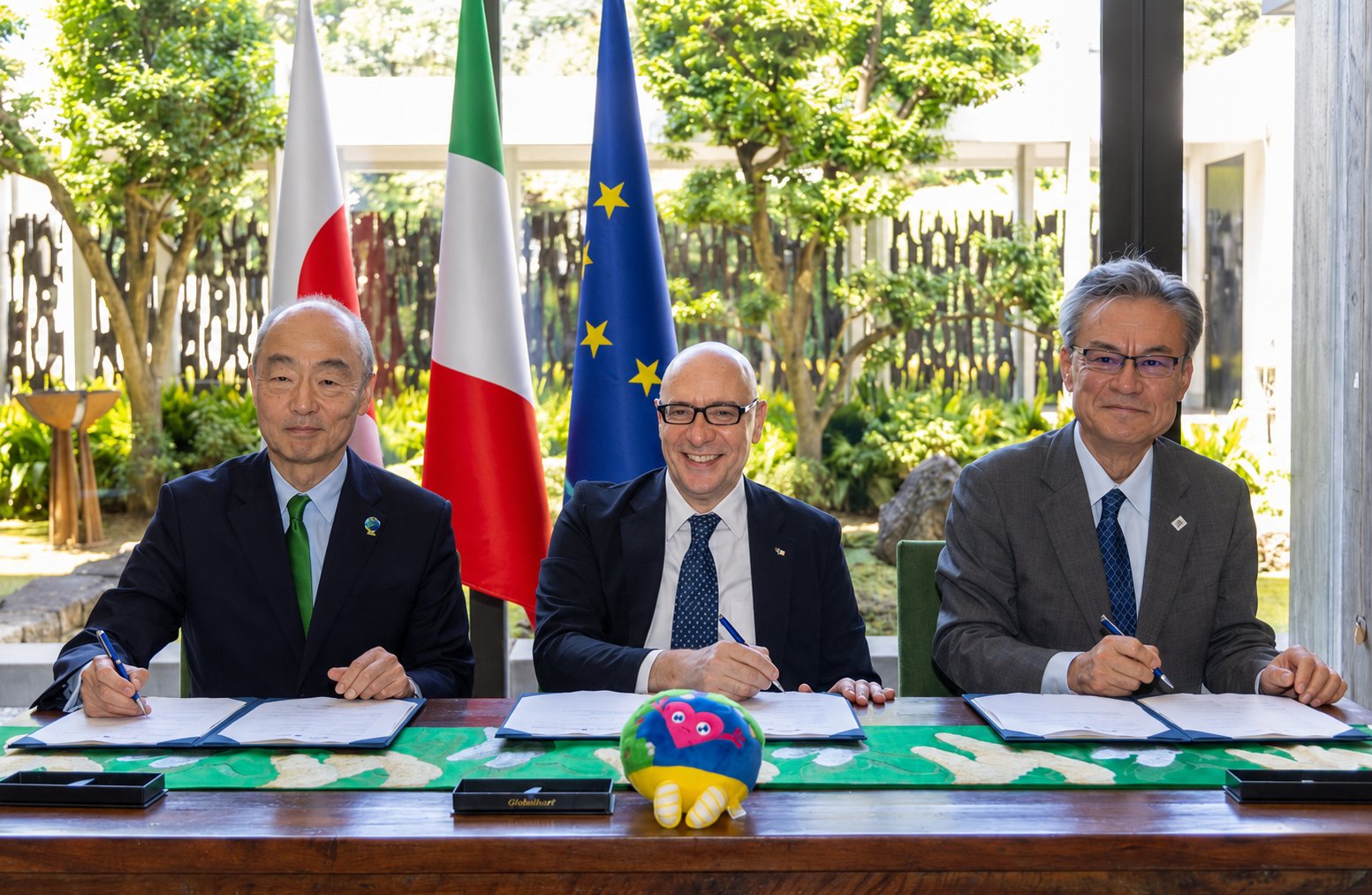 イタリアとの公式参加契約の調印式。日本・イタリア・EUの旗の前で3人が契約書に署名し、机にトゥンクトゥンクの人形が置かれている
