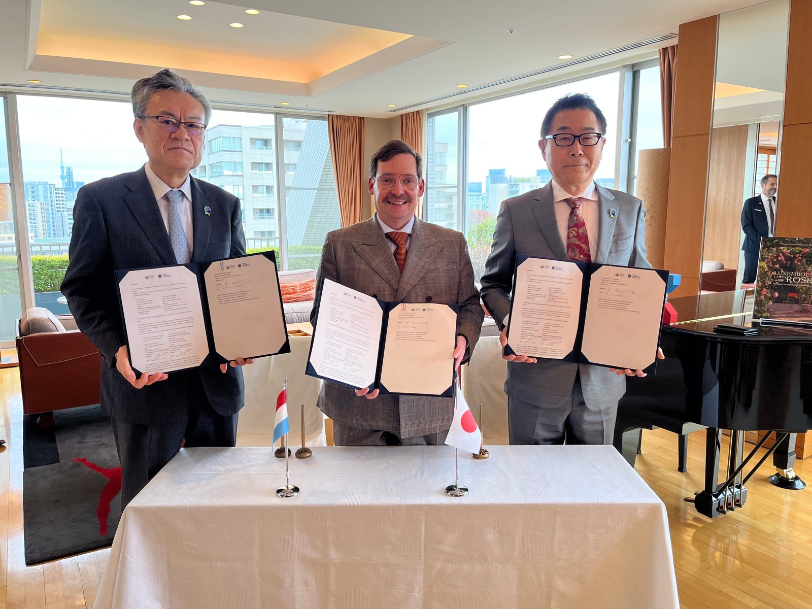 ルクセンブルクとの公式参加契約の調印式。3人が署名した契約書を掲げ、机にルクセンブルクと日本の小旗が置かれている