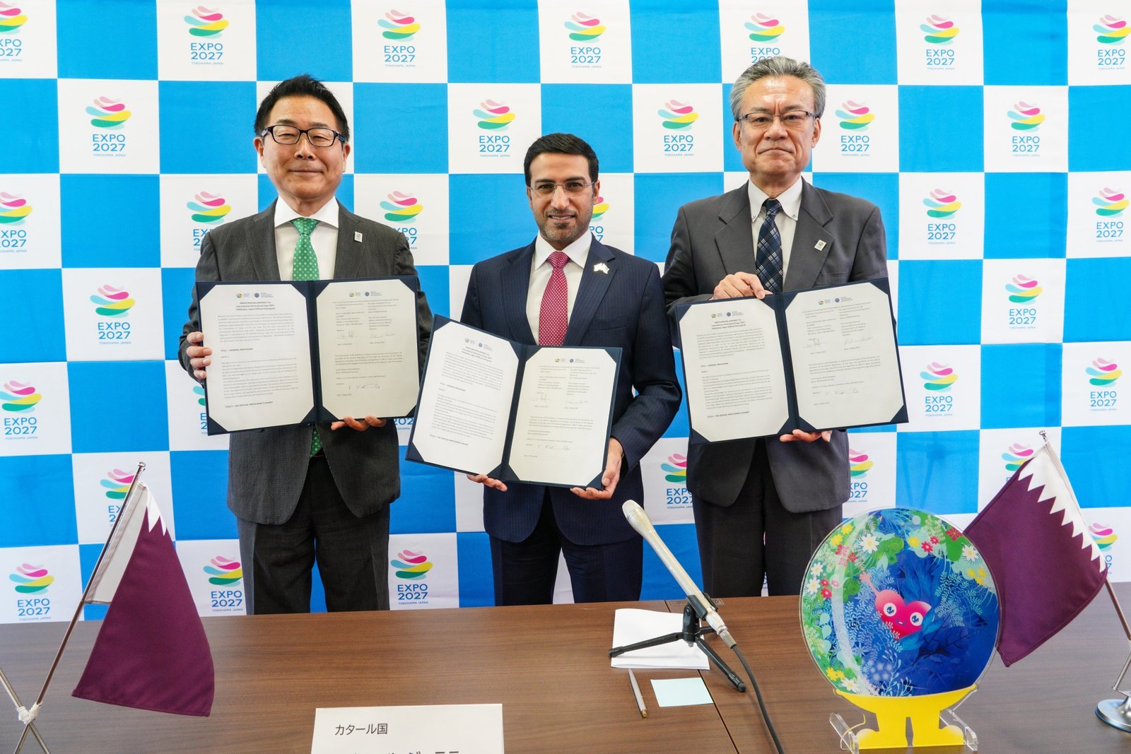 カタール国との公式参加契約の調印の様子。EXPO 2027のロゴのパネルの前で3人が契約書を掲げ、机にカタールと日本の旗とトゥンクトゥンクの人形
