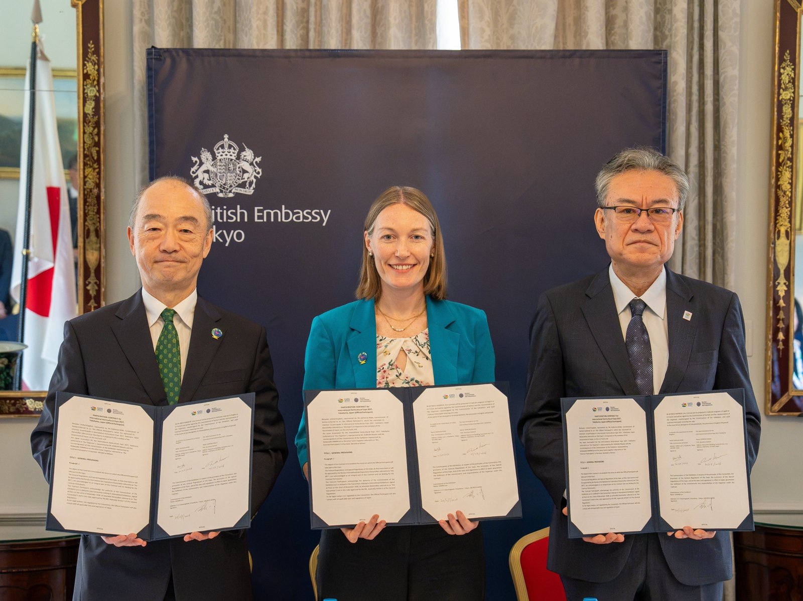 駐日英国大使館での公式参加契約の調印式。3人が署名した契約書を掲げている