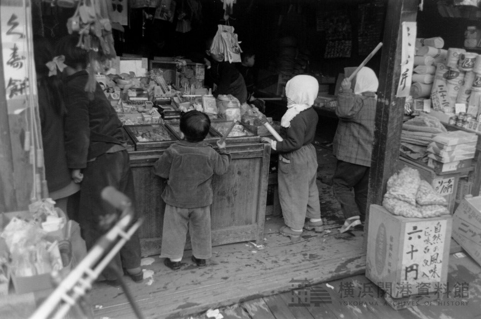 昭和30年頃の駄菓子屋の店先で買い物をする子どもたち3人の後ろ姿。棚に菓子や日用品が並ぶ